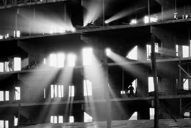 imagem da construção de Brasília, com a luz do sol penetrando por espaços do prédio e contrastando com ele e figuras humanas
